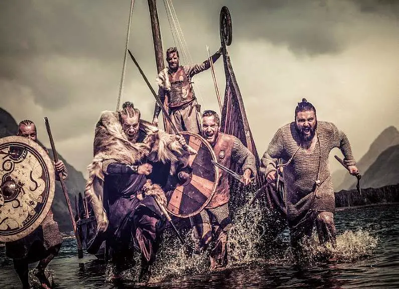 A small group of vikings running angrily with weapons out of a boat in water