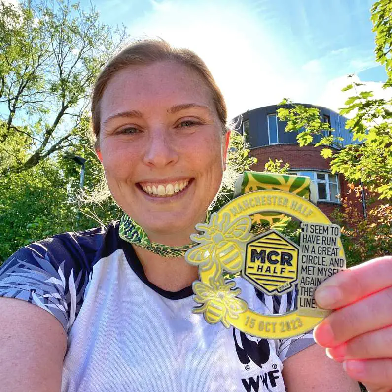 Mel holding up a Manchester Half medal, smiling in the sunshine after finishing the race