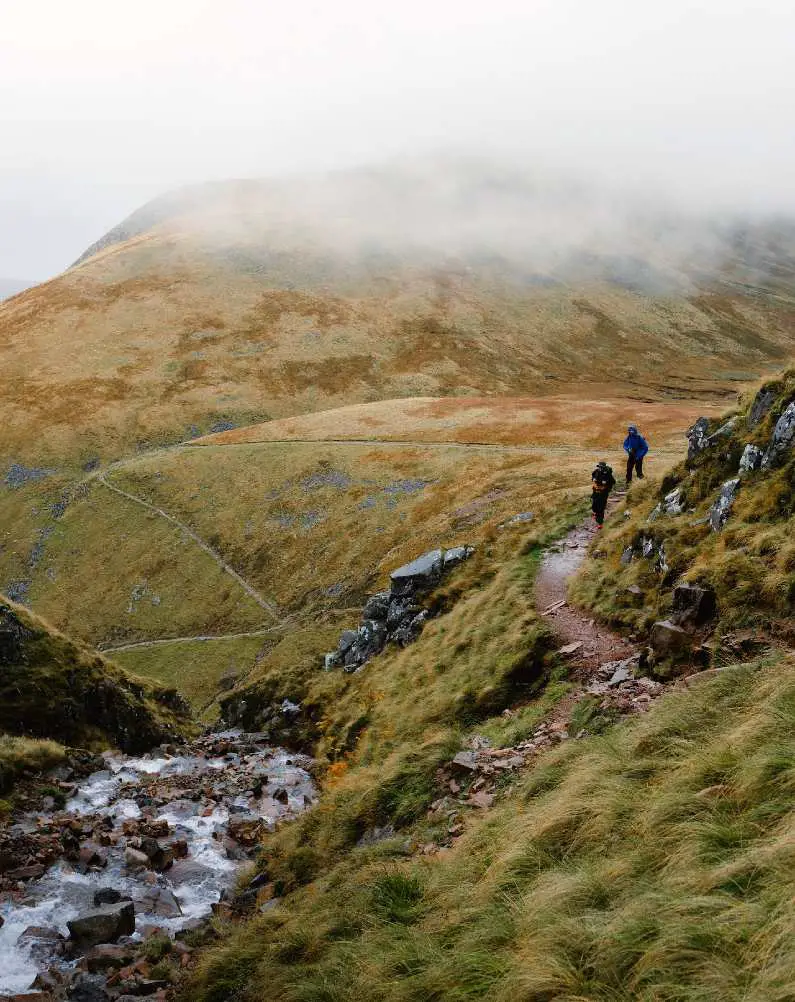 Two hikers in the distant on a path on the way up to Ben Nevis with heavy mist and a stream running 