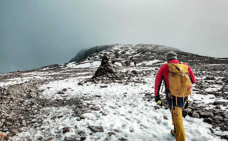Hiker wearing yellow and red hiking clothes walking across the summit of Ben Nevis covered in snow