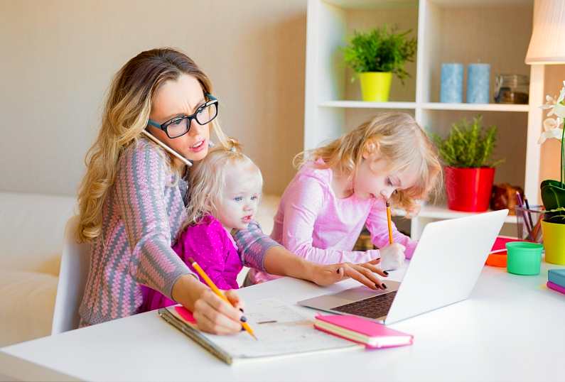 Young mum sat at a white desk with her two little girls sat around her as she is stressed and trying to work at her laptop