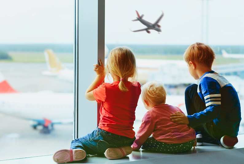 Three small, blonde children looking out the window at an airport watching plane fly in